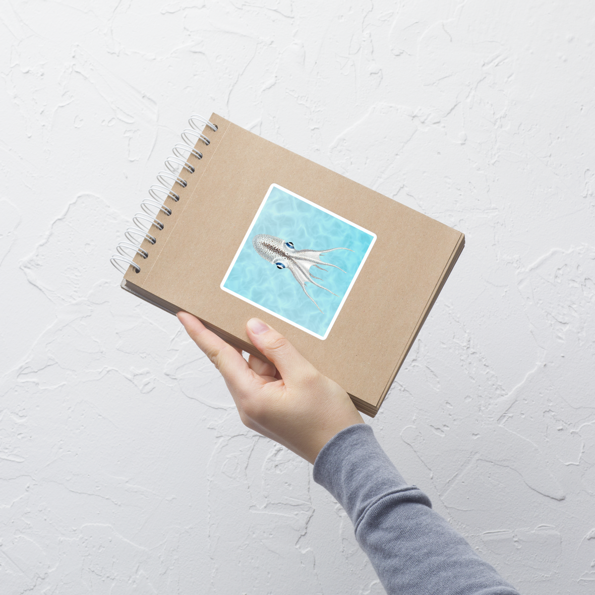 Hand holding a spiral-bound notebook with a cover illustration of an octopus on a light gray background