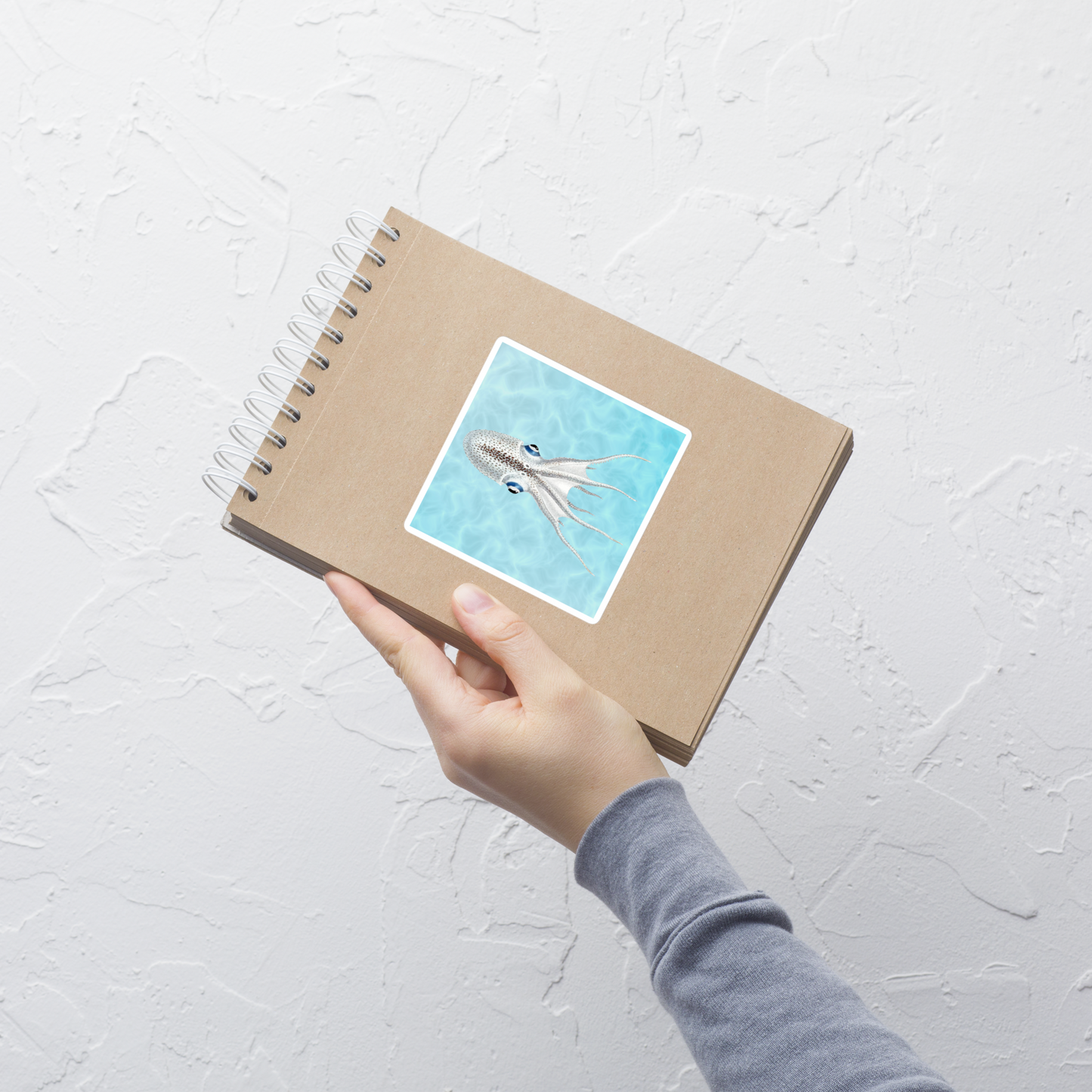 Hand holding a spiral-bound notebook with a cover illustration of an octopus on a light gray background