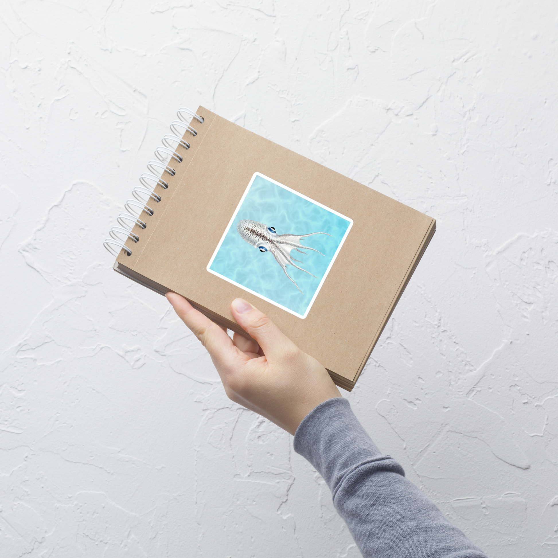 Hand holding a spiral-bound notebook with a cover illustration of an octopus on a light gray background