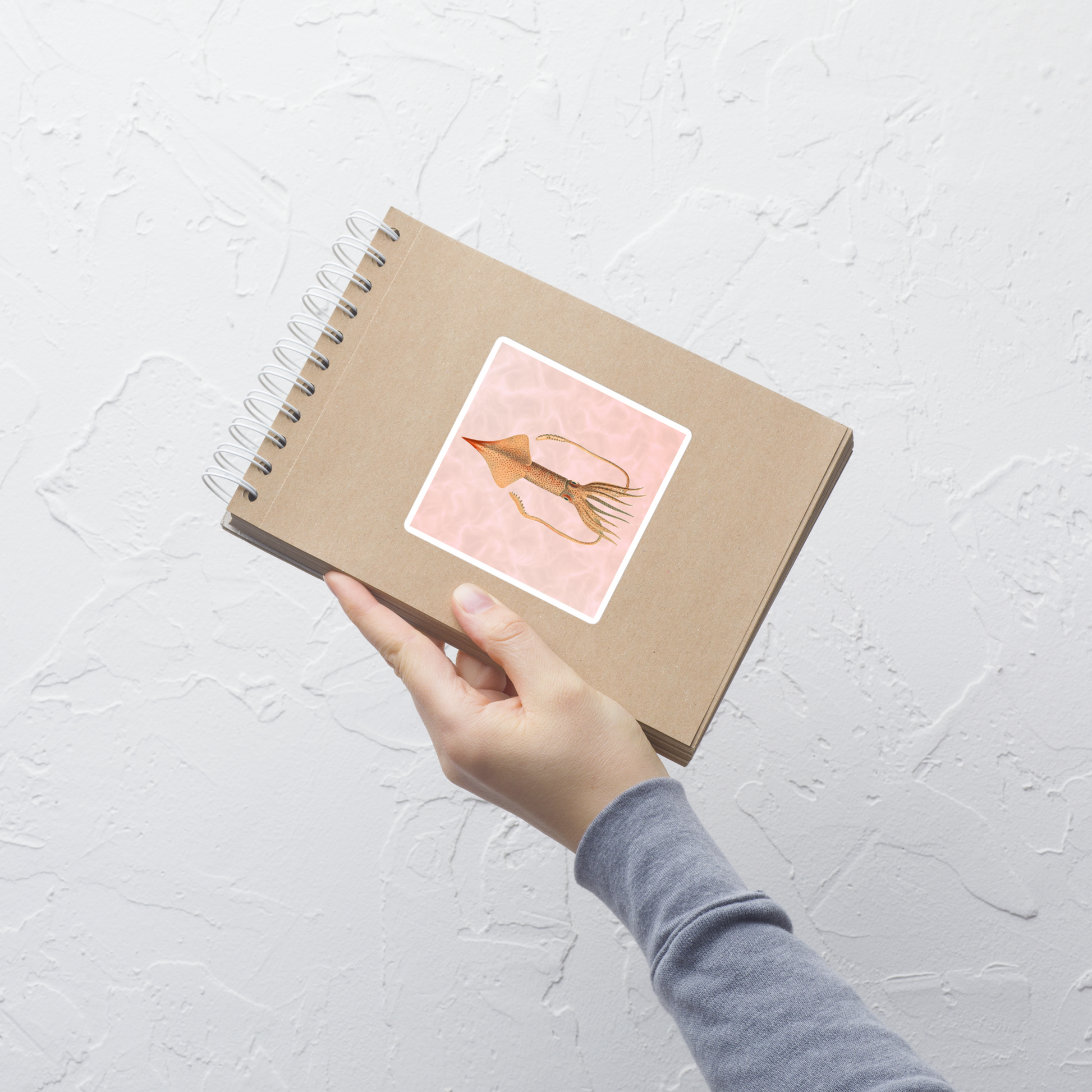 Hand holding a brown notebook with Angel Clubhook Squid 4x4 vinyl sticker with red background 