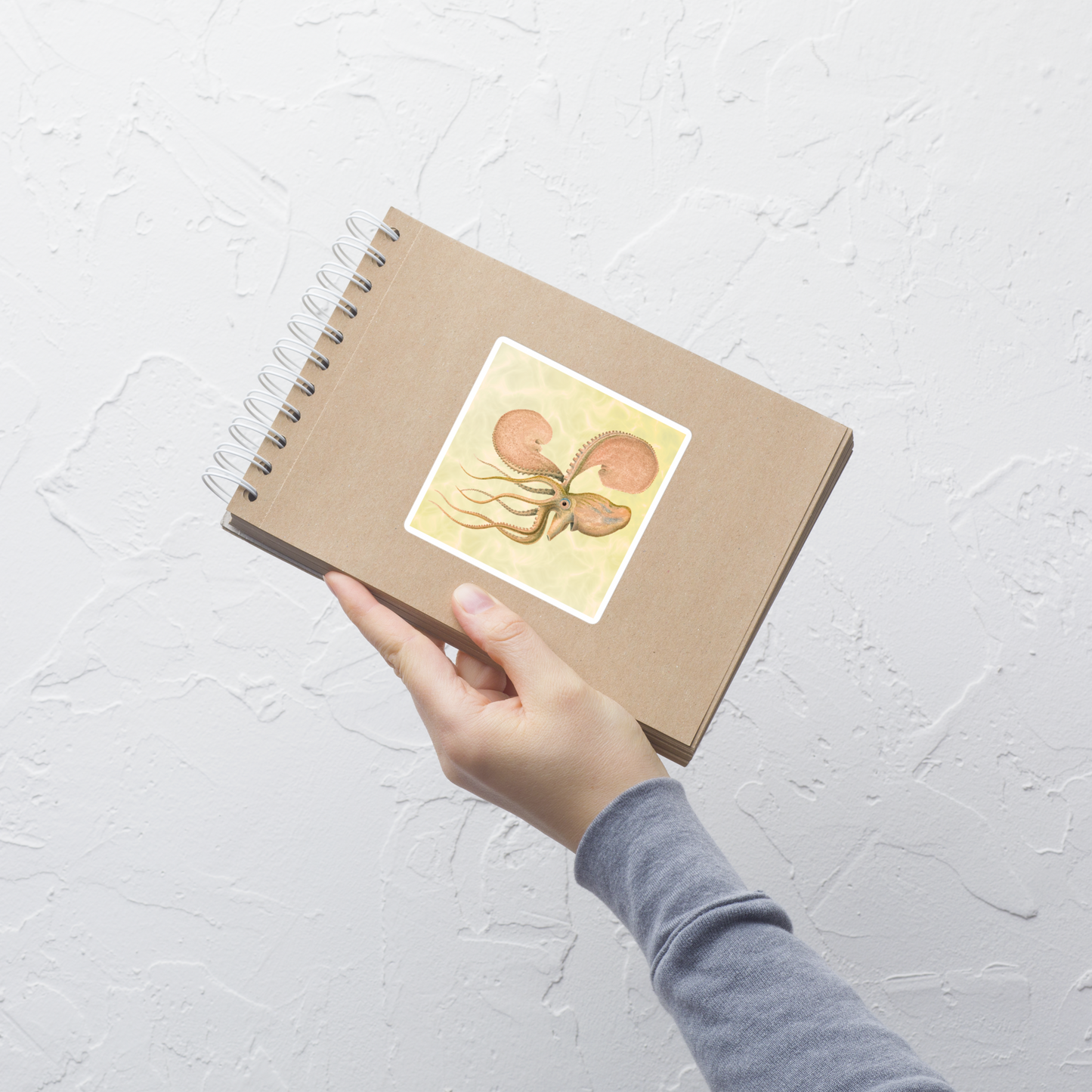 Hand holding a brown notebook with an Argonauta Argo paper nautilus 4x4 vinyl sticker with a yellow background 
