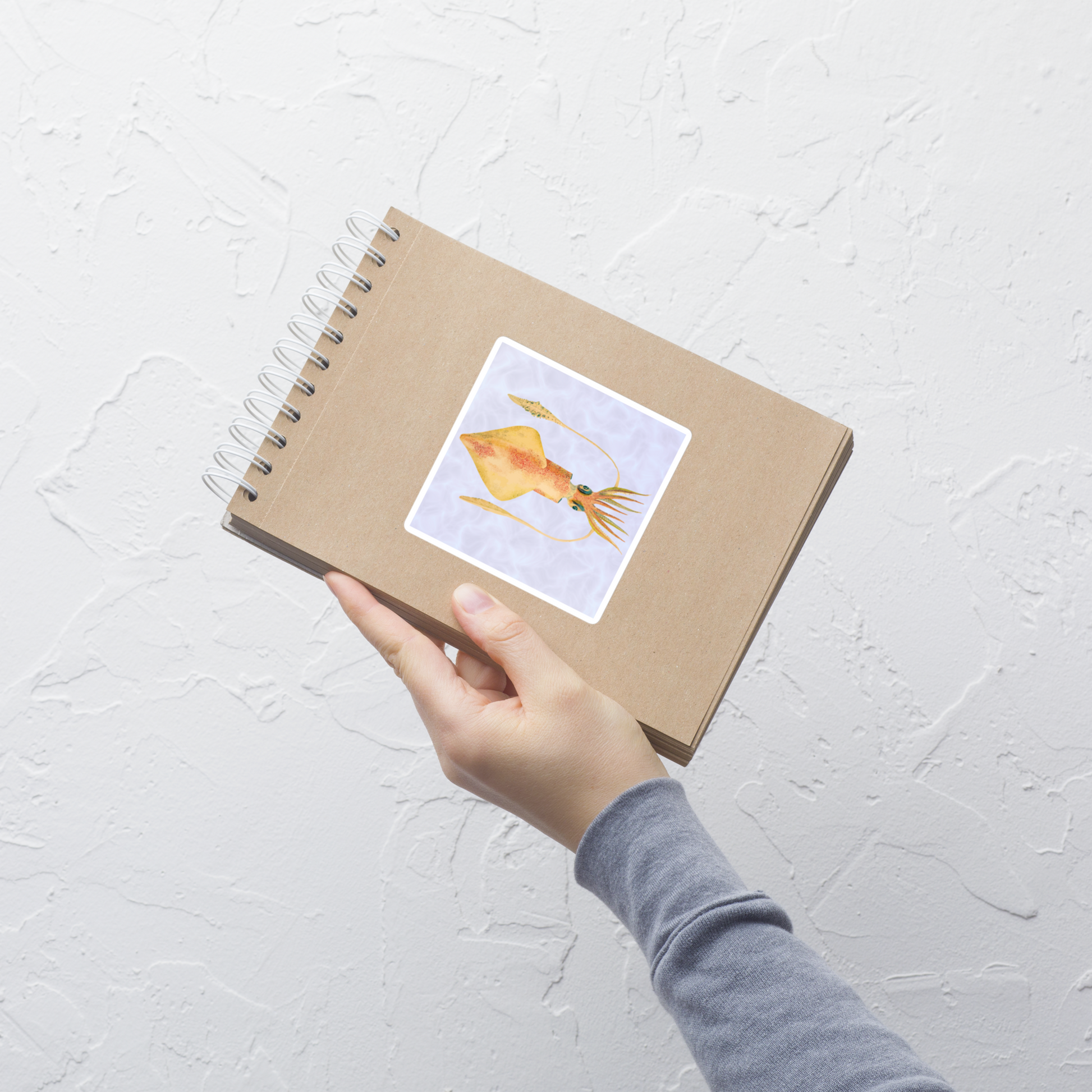 Hand holding a spiral-bound notebook with a Loligo Vulgaris squid 4x4 vinyl sticker with purple background 