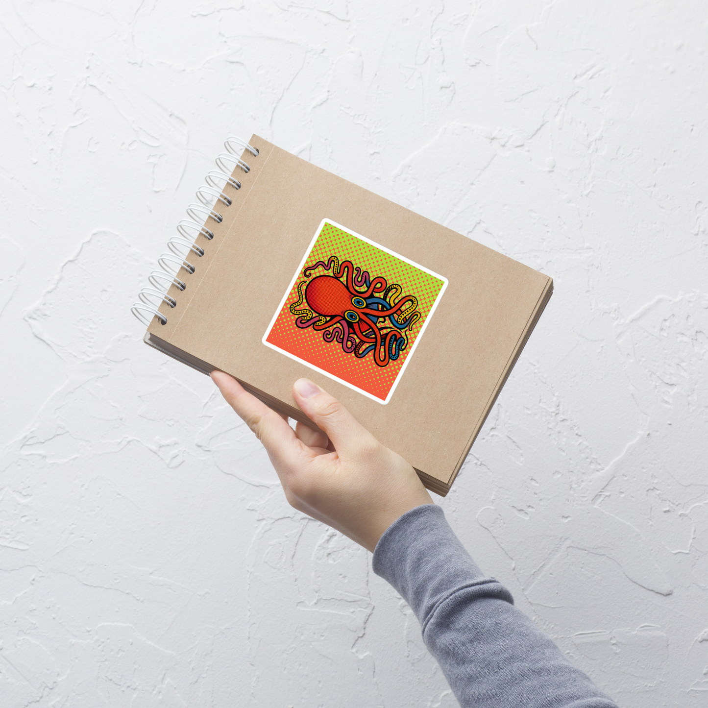 Hand holding a spiral-bound notebook with 8-Armed Cuttlefish pop art vinyl sticker 4x4 — red, blue, and orange cuttlefish on halftone background