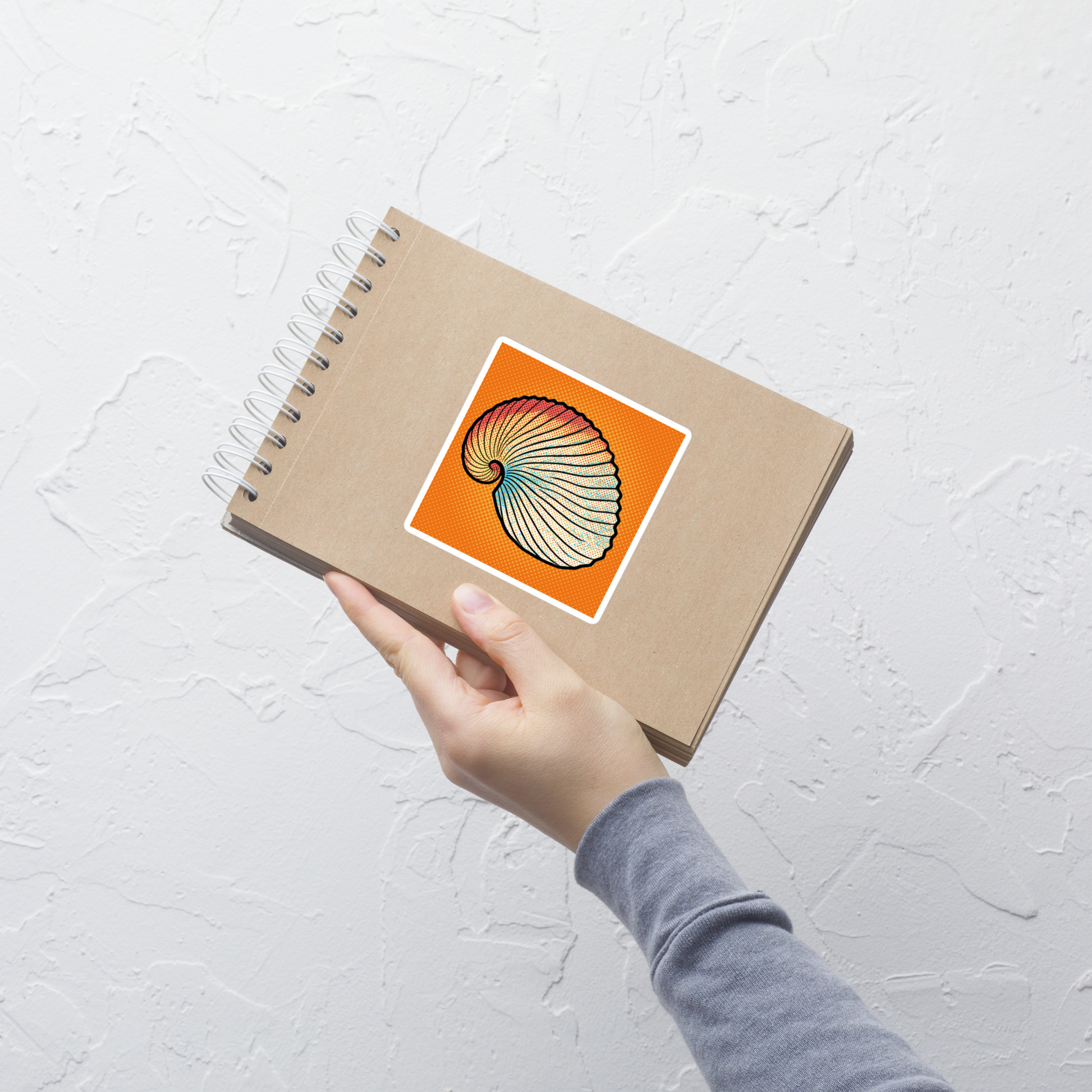 Hand holding a spiral-bound notebook with Argo Shell pop art vinyl sticker 4x4 — argonaut paper nautilus shell on orange halftone background