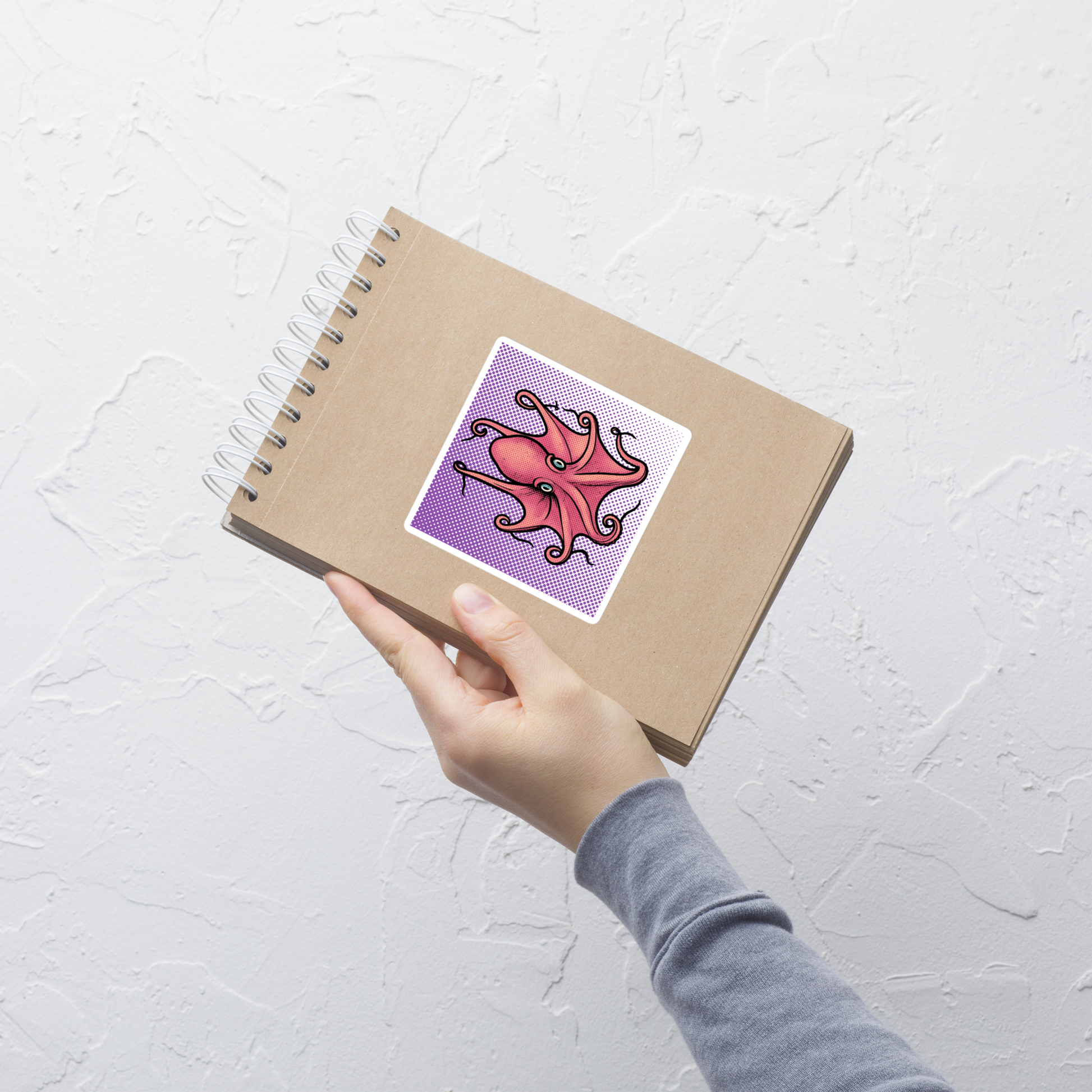Hand holding a spiral-bound notebook with Eledon Aldrovandi Octopus pop art vinyl sticker 4x4 — pink octopus on purple halftone background