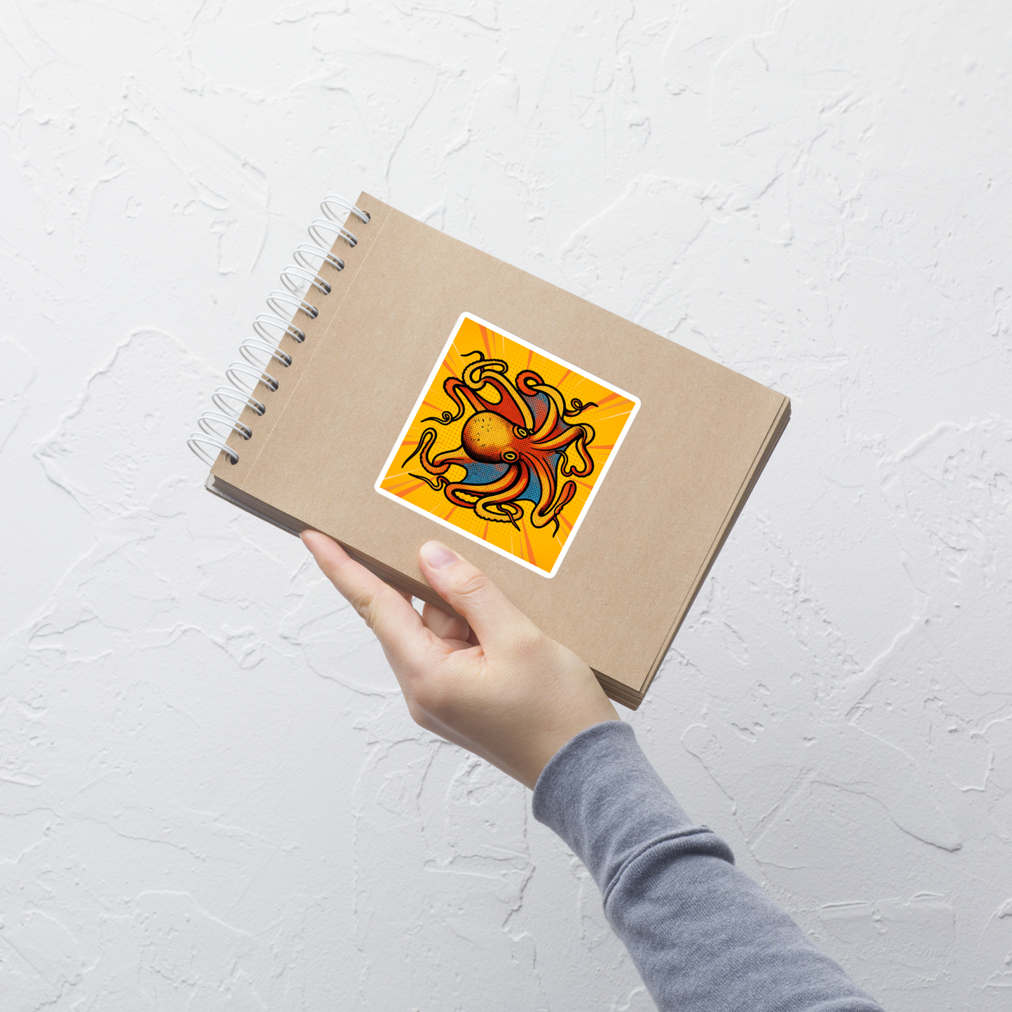 Hand holding a spiral-bound notebook with Octopus Vulgaris pop art vinyl sticker 4x4 — blue and orange octopus on yellow sunburst background