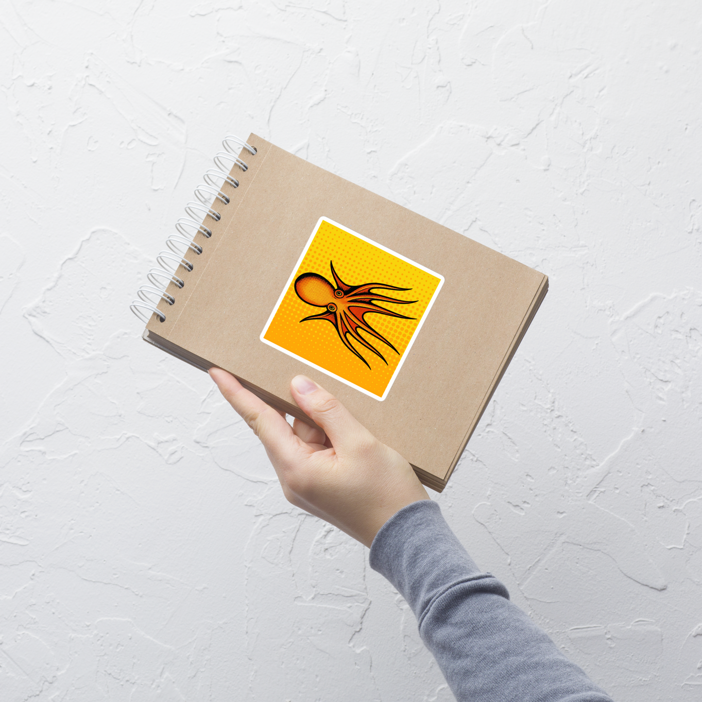 Hand holding a spiral-bound notebook with a Four Horn Tetracirrhus Octopus pop art vinyl sticker 4x4 — orange octopus with four horns on a yellow background