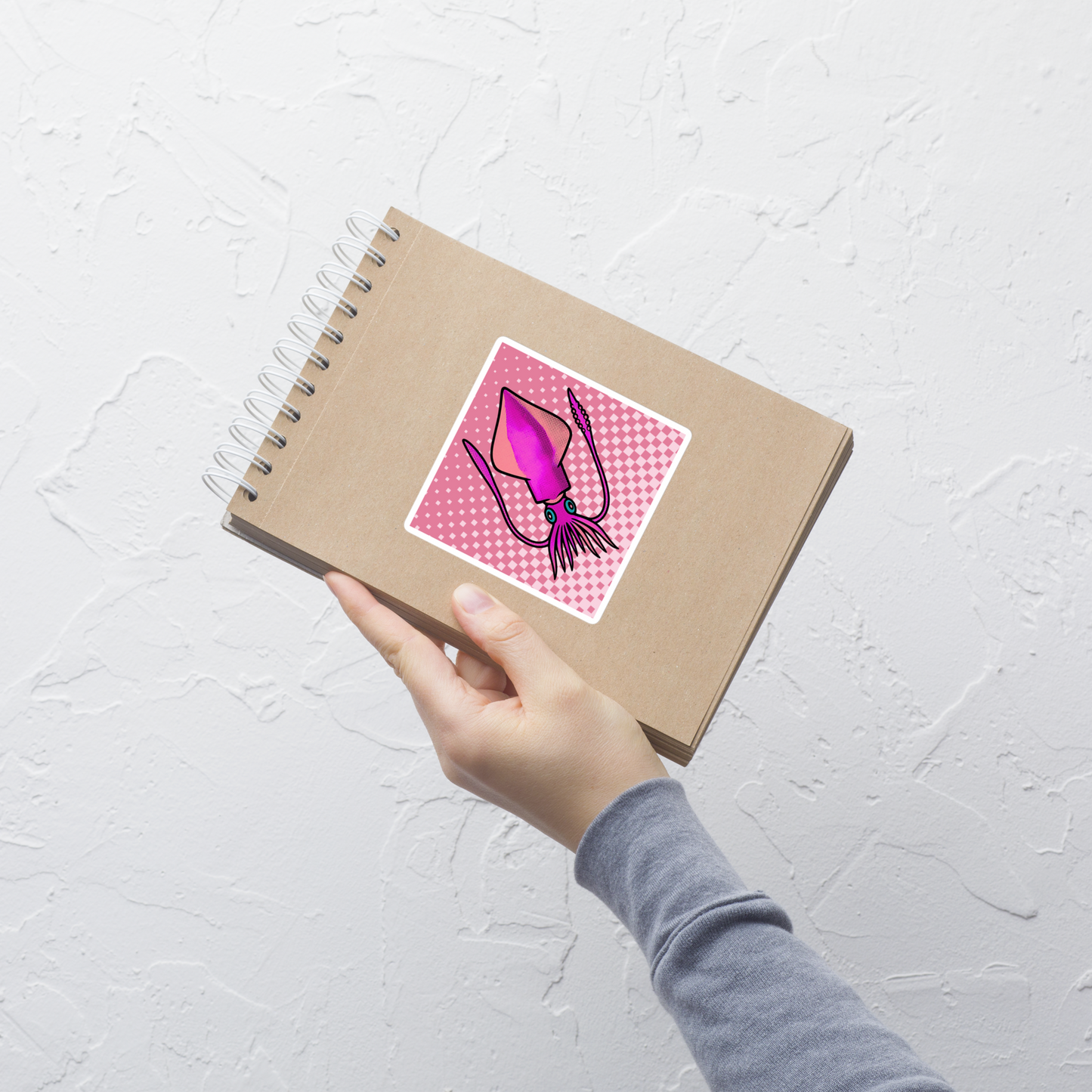 Notebook with Loligo Vulgaris Squid pop art vinyl sticker 4x4 — pink and magenta squid on a checkered pink background, held by a hand on a light gray background