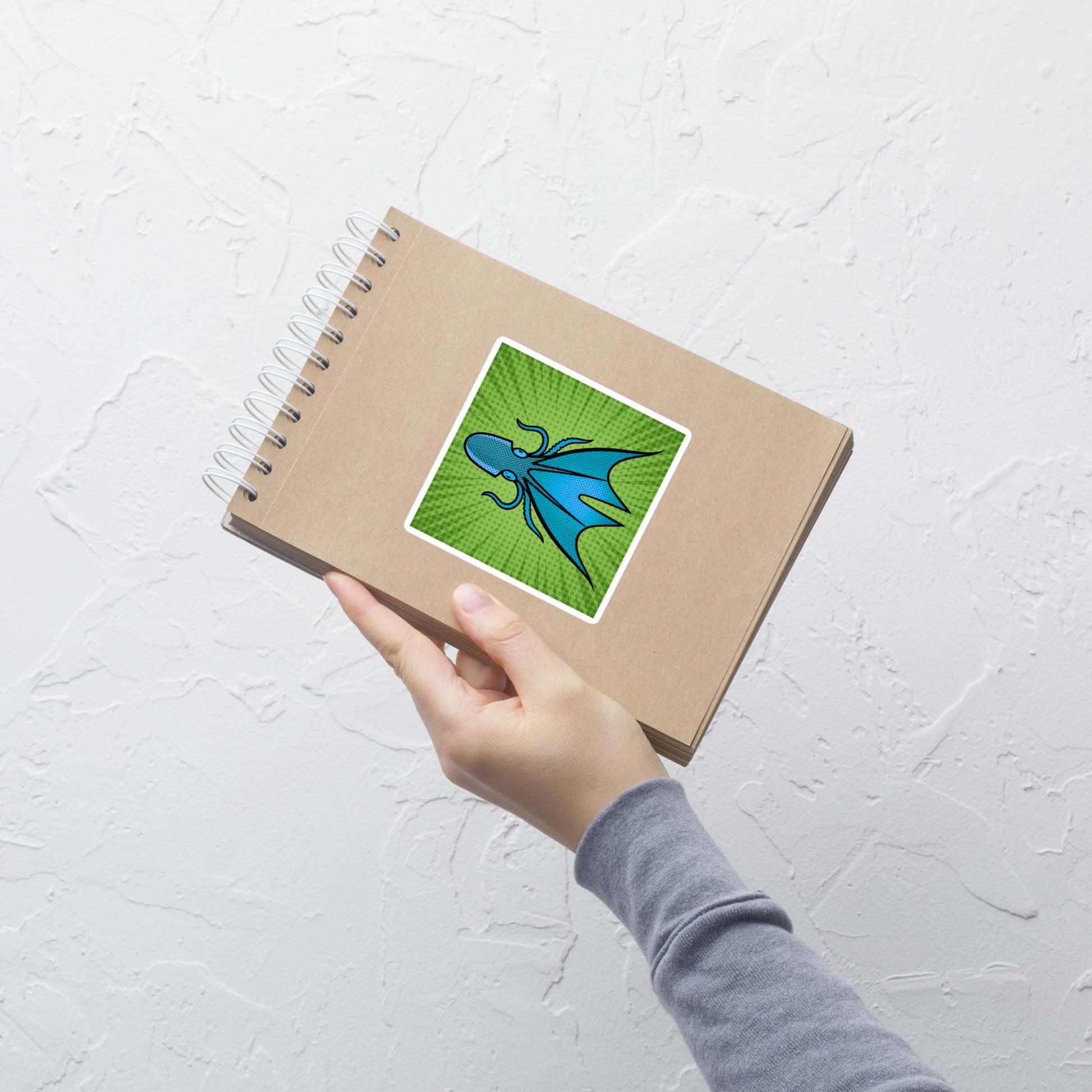 Hand holding a spiral-bound notebook with a Common Blanket Octopus pop art vinyl sticker 4x4 — blue and teal octopus with webbing on green sunburst background on a light gray background