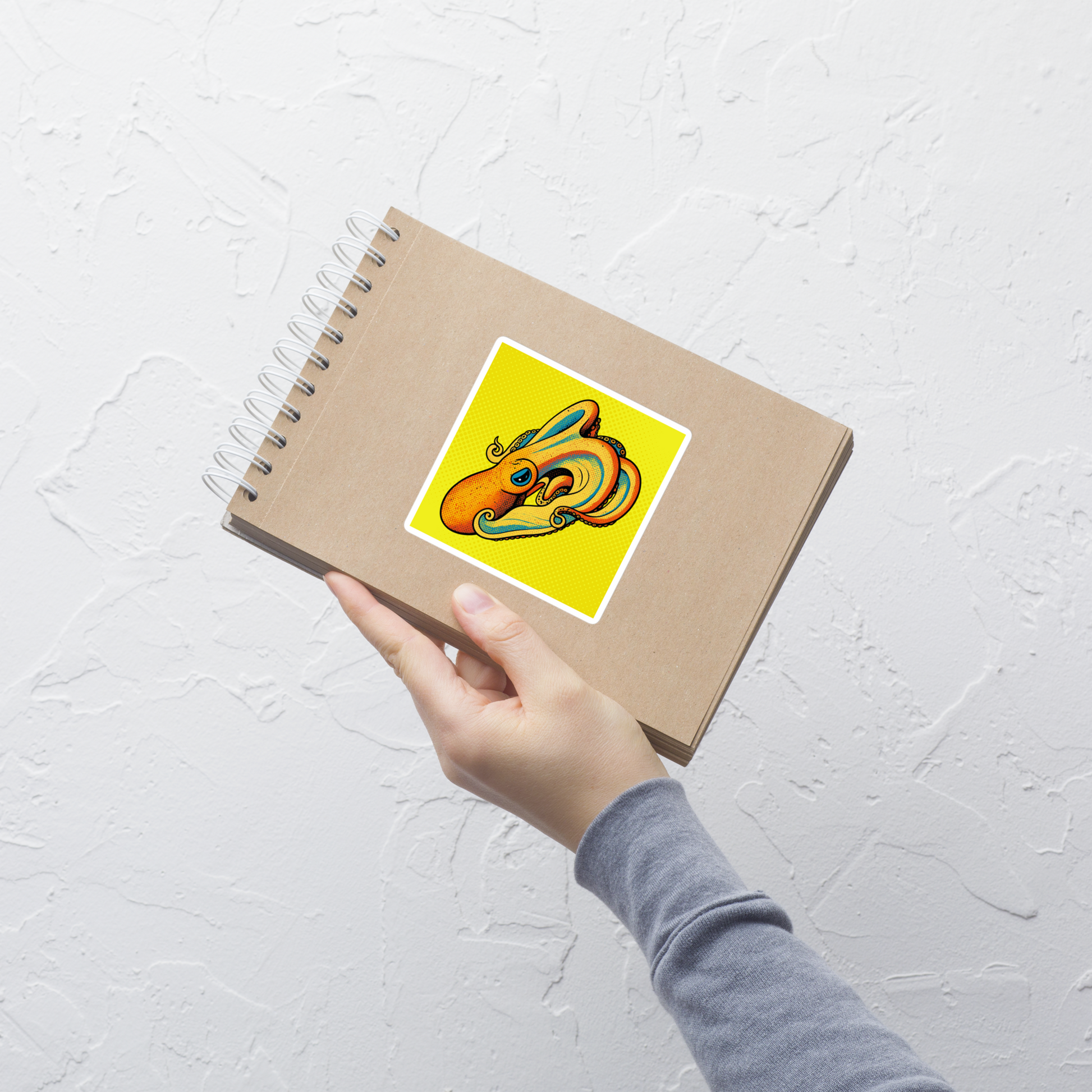 Notebook with a Velodona Octopus pop art vinyl sticker 4x4 — orange and teal octopus on bright yellow halftone background on a white textured background