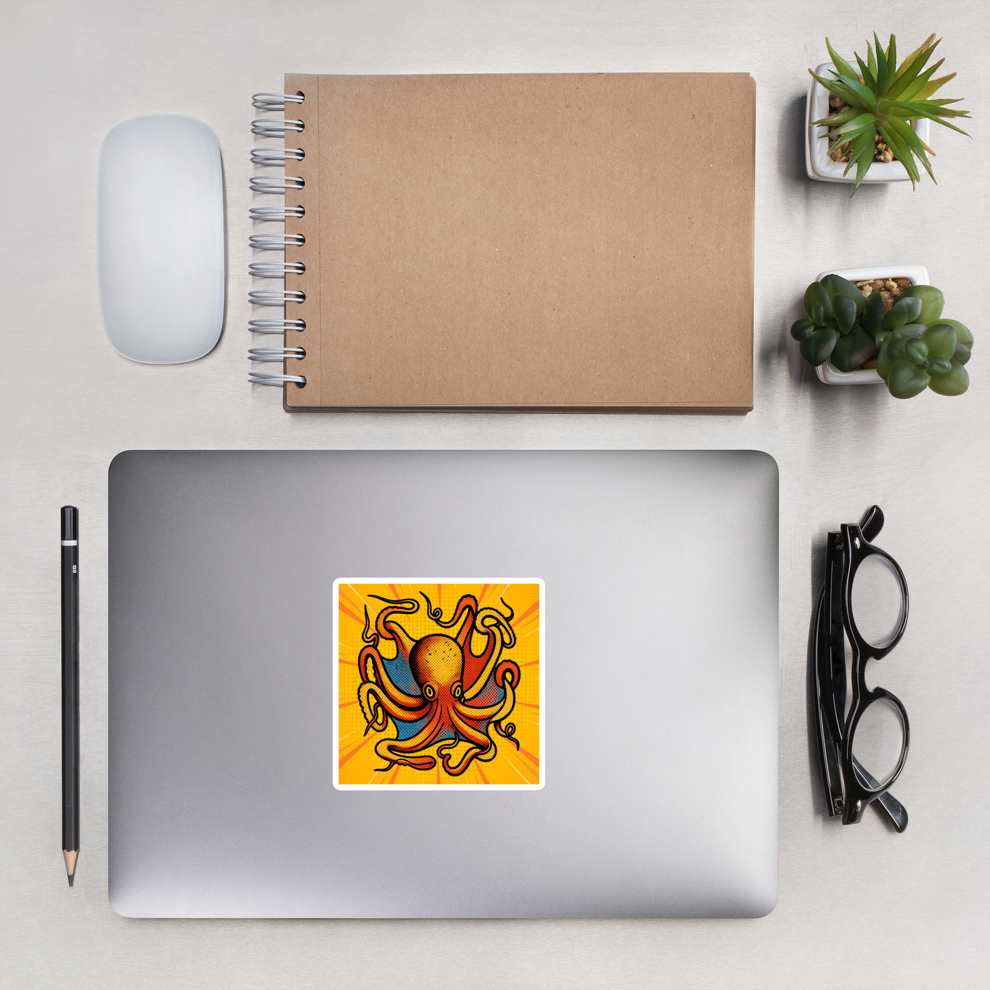 Laptop with Octopus Vulgaris pop art vinyl sticker 4x4 — blue and orange octopus on yellow sunburst background on a desk with a notebook, mouse, pencil, glasses, and plants.