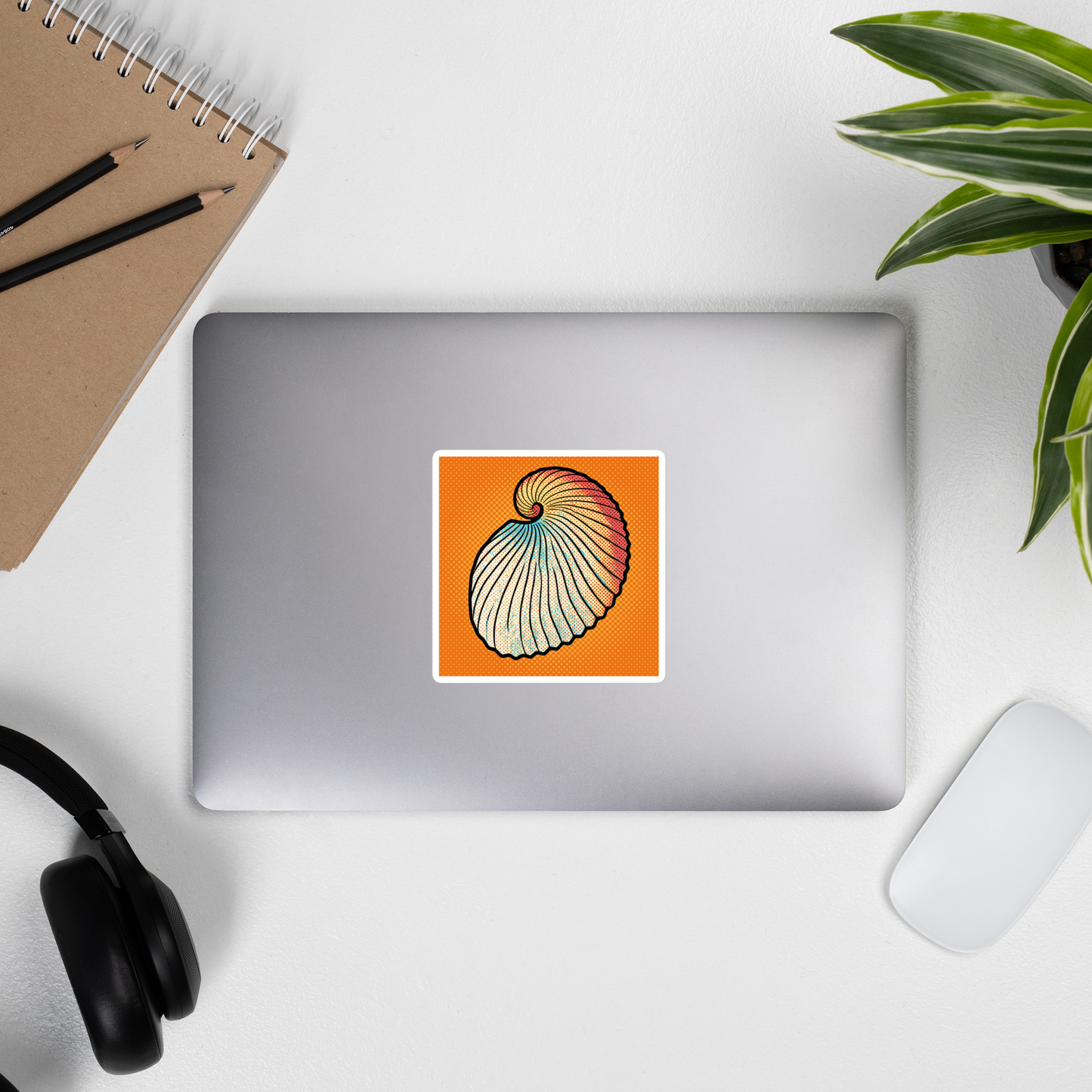Laptop with Argo Shell pop art vinyl sticker 4x4 — argonaut paper nautilus shell on orange halftone background on a desk with a notebook, pencils, and headphones.