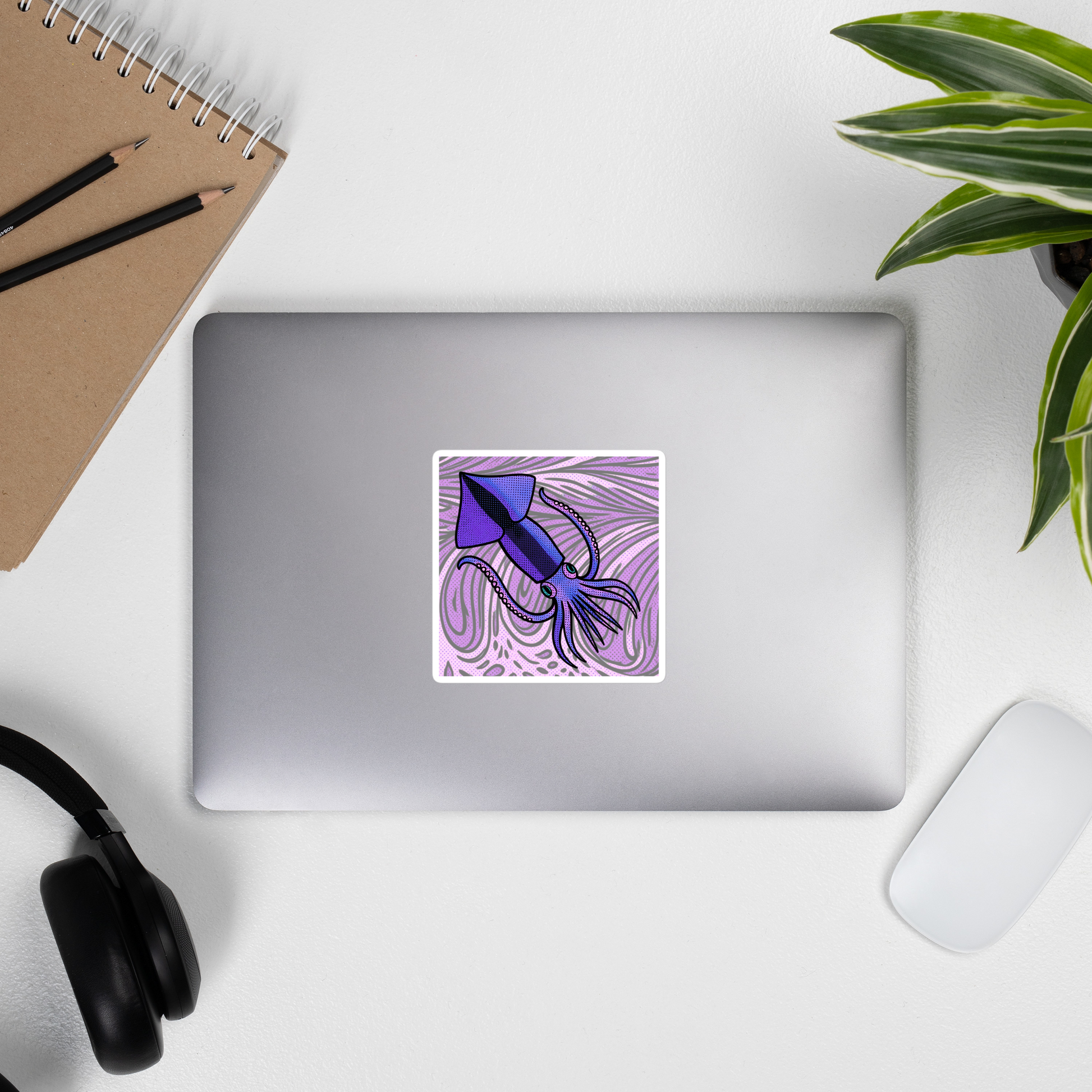 Laptop with a Loligo Todarus Squid pop art vinyl sticker, 4x4 — purple squid on a lavender swirl halftone background on a white surface, with a plant and a notebook.