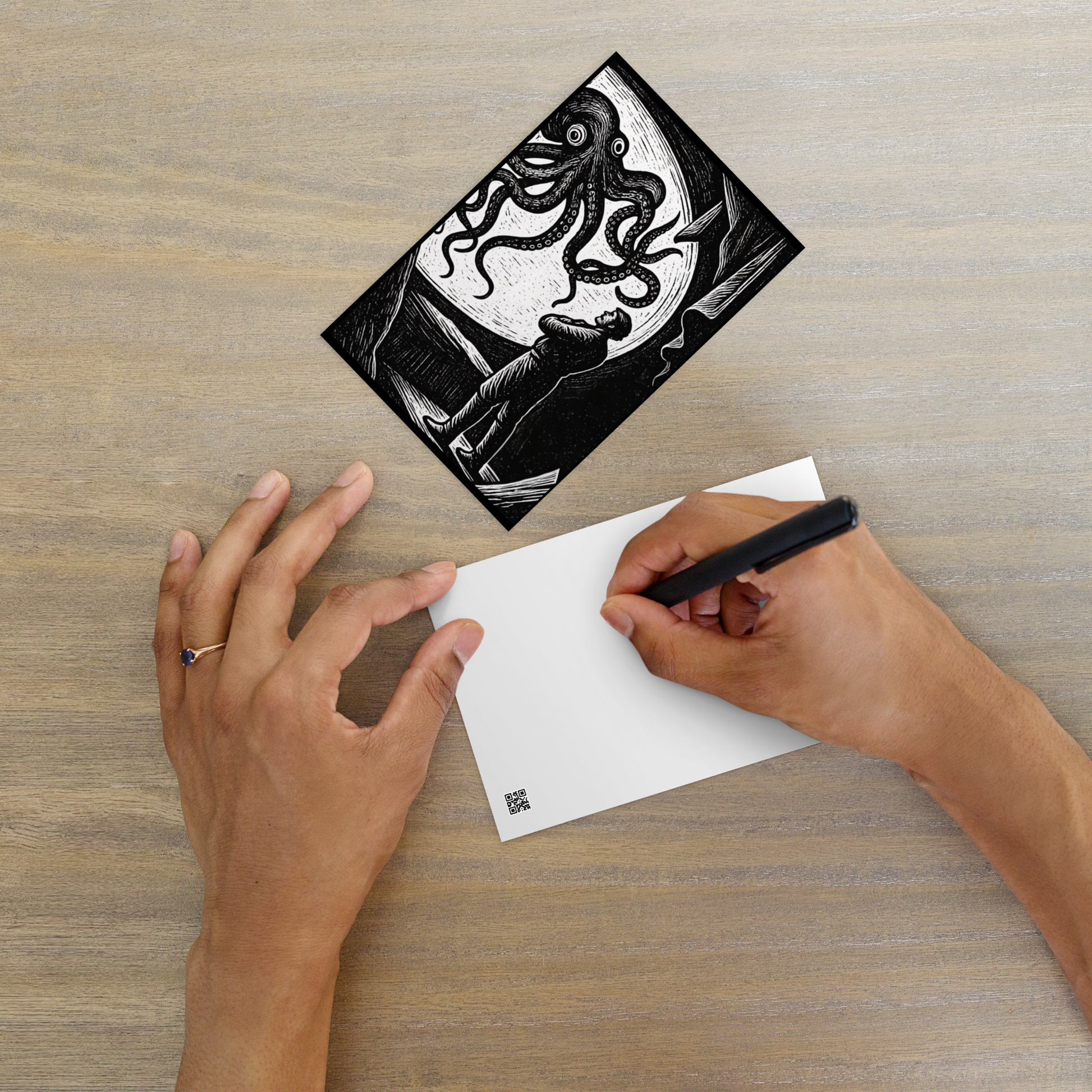 Person writing on a white card with a black pen, next to a 20,000 Leagues Under the Sea lithocut postcard — black and white giant squid scene from Jules Verne illustration