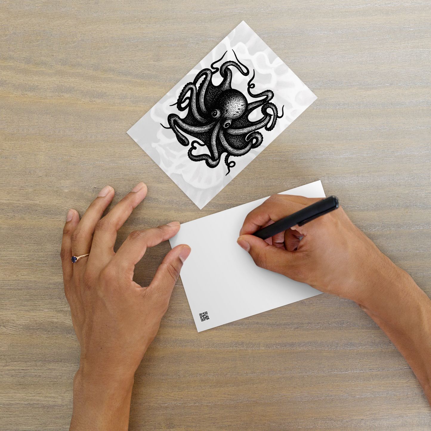 Person holding a black pen over a white card with a Octopus Vulgaris lithocut postcard — black and white illustration of common octopus with radiating arms