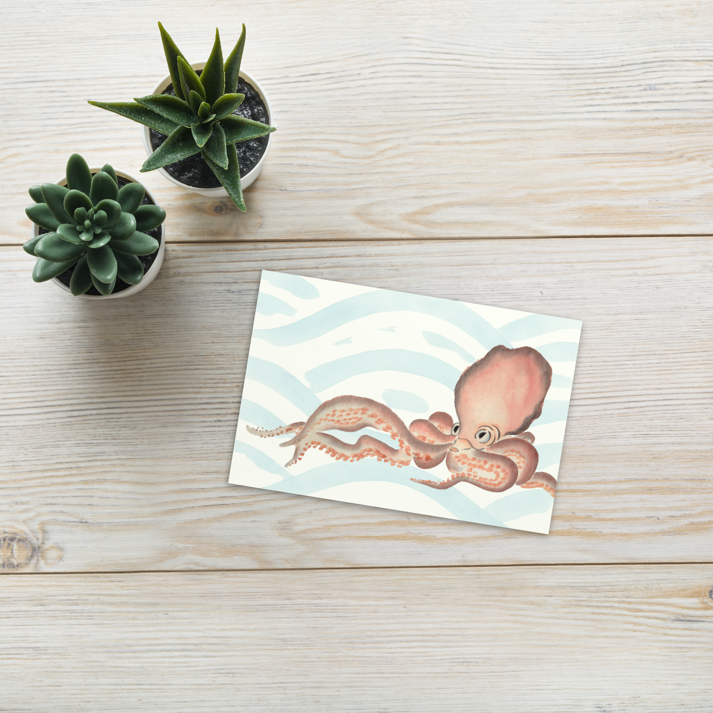 Note card with an Japanese octopus illustration postcard 4x6 — soft pink watercolor octopus with blue ocean wave background on premium cardstock on a wooden surface with two small potted plants.